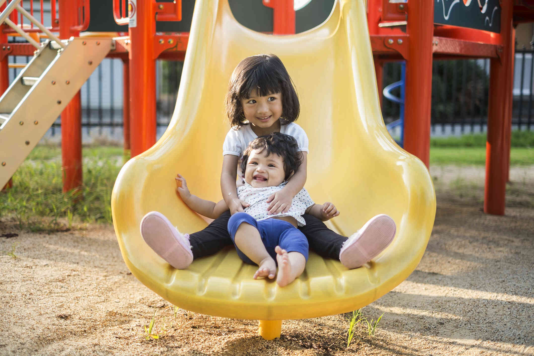 A little girl and her toddler sibling slide down a slide together while smiling.